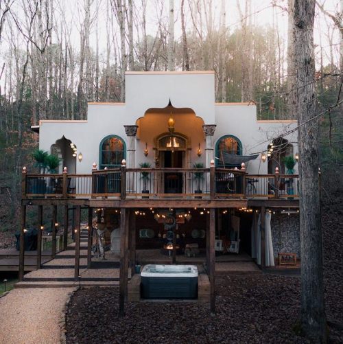 A charming, elevated house surrounded by trees, featuring a spacious deck, hot tub, and decorative lights.