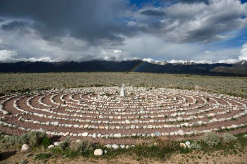 A stone labyrinth set in a desert landscape, surrounded by mountains and dramatic clouds, with a rainbow in the distance.