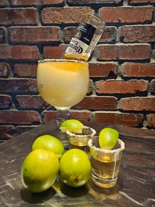 A margarita glass with a bottle upside down, surrounded by limes and shot glasses on a metal surface against a brick wall.