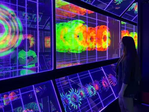 A person observes vibrant, glowing patterns in a dark room with neon lights and interactive displays.