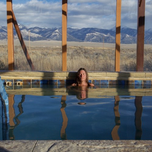 A person relaxes in a pool with mountains and a cloudy sky in the background, reflecting in the water.