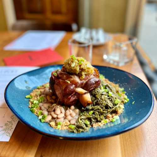 A plate featuring a large piece of meat with greens, beans, and garnishes, set on a wooden table.