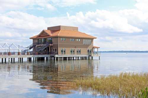 A modern building with a red roof sits on stilts over calm water, reflecting in the lake under a cloudy sky.