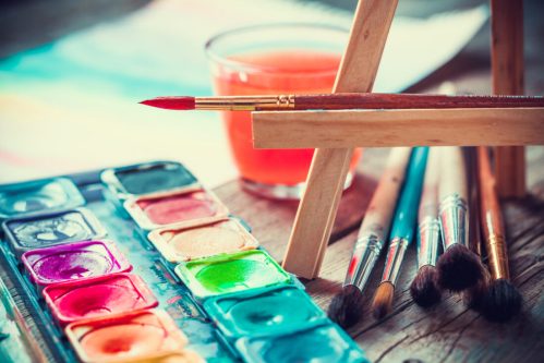 A watercolor palette, brushes, and a glass of orange liquid on a wooden table, with an easel in the background.