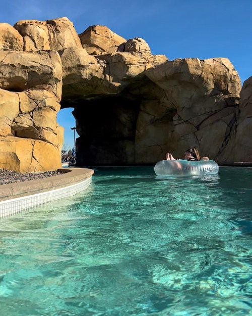 A person relaxes on a clear inflatable float in a turquoise pool, surrounded by rocky formations and blue skies.