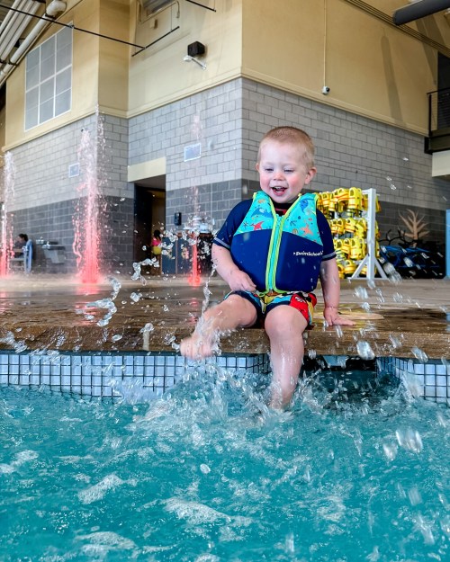 A joyful child in a life jacket splashes in a pool, sitting on the edge with water spraying around.