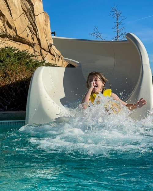 A child in a yellow life jacket slides down a water slide, splashing into a pool on a sunny day.