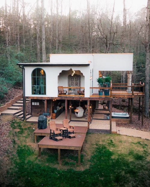 A modern treehouse with a spacious deck, surrounded by trees and featuring outdoor seating and a hot tub.