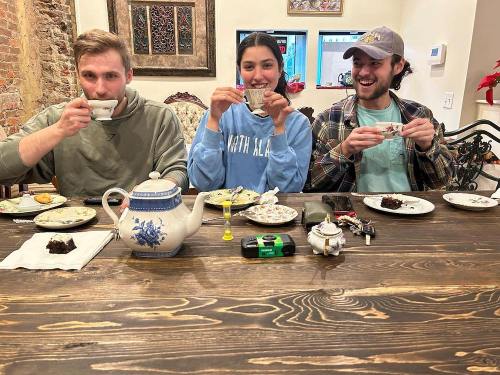 Three friends sit at a table, smiling and holding teacups, with a teapot and desserts in front of them.