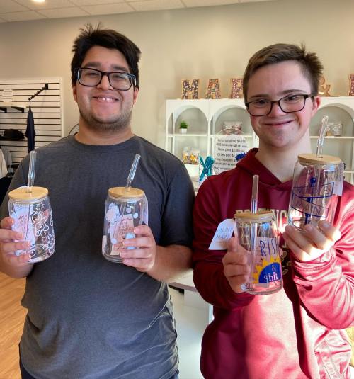 Two young men hold decorated glass jars with wooden lids and straws, smiling in a bright, cheerful store setting.