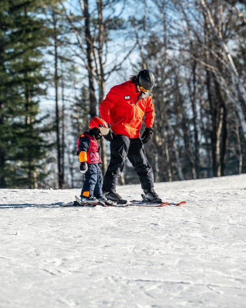 A parent teaches a child to ski on a snowy slope, surrounded by trees and a clear blue sky.