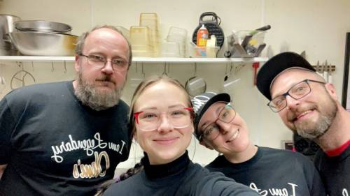 A group of four people smiling in a kitchen, wearing matching black shirts with a playful design.