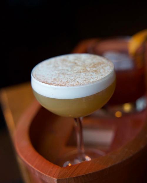 A frothy cocktail in a coupe glass, topped with a dusting of cinnamon, sits on a wooden surface.