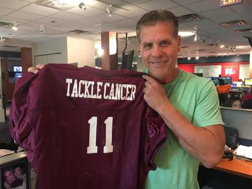 A man holds up a maroon football jersey with "TACKLE CANCER" and the number "11" on the back, smiling in an office setting.