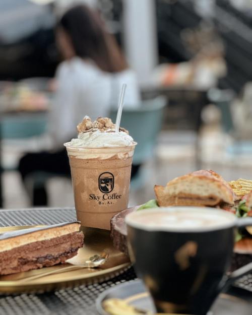 A frosty coffee drink topped with whipped cream and snacks on a table, with a blurred person in the background.