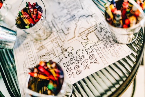 Colorful crayons in metal cups beside a coloring page for "Alley Cats Coffee Bar" on a glass table.