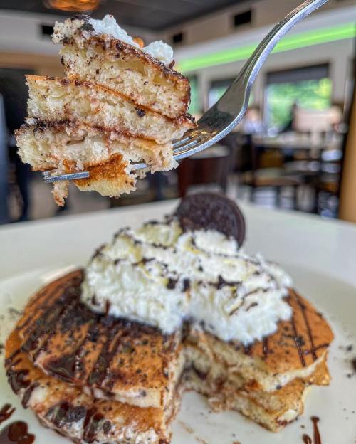 A fork holding a piece of stacked pancakes topped with whipped cream, chocolate drizzle, and an Oreo cookie.
