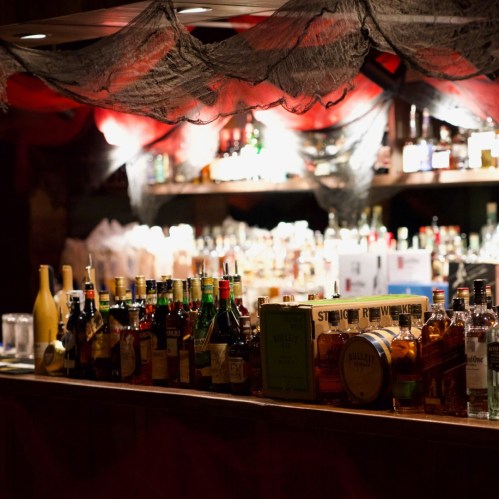 A dimly lit bar with a variety of liquor bottles lined up on the counter, decorated with cobwebs and red fabric.