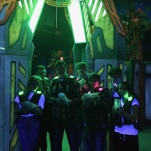 A group of six people poses under neon lights, holding laser tag equipment in a vibrant, themed arena.