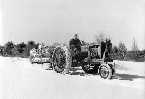 A man drives a tractor pulling a log sled through a snowy landscape. Trees are visible in the background.