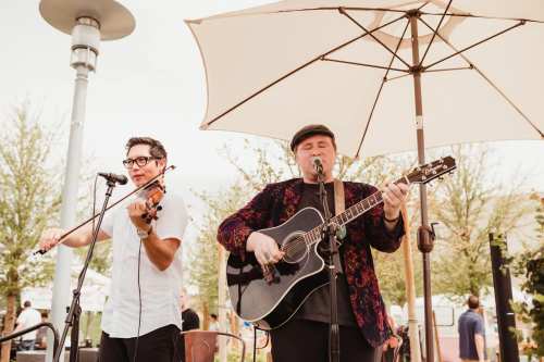 Two musicians perform outdoors under an umbrella; one plays guitar and sings, while the other plays the violin.