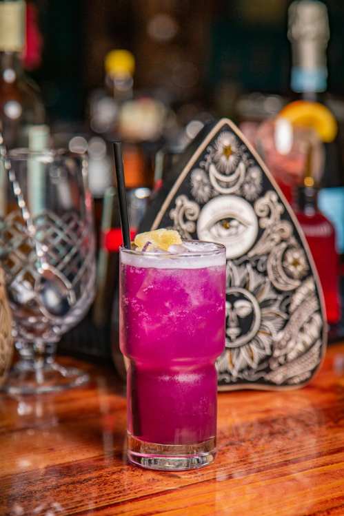 A vibrant purple cocktail with a lemon slice and straw, set against a blurred bar background.