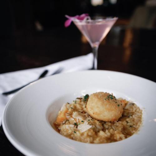 A plate of creamy risotto topped with a golden fried ball, accompanied by a pink cocktail in a glass.