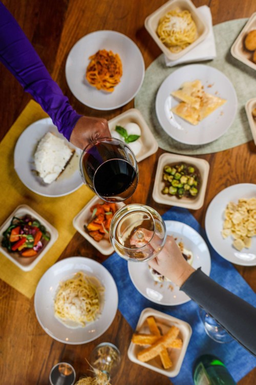 A table filled with various dishes and two hands clinking glasses of wine above the spread.