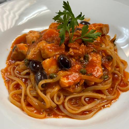 A plate of pasta topped with a rich tomato sauce, olives, capers, and garnished with a sprig of parsley.