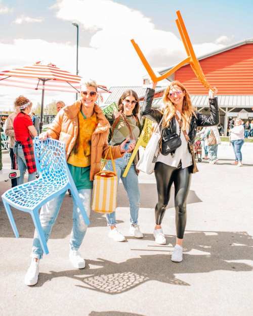 Three women joyfully walk outdoors, each holding colorful chairs and bags, with a lively market scene in the background.