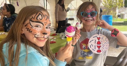 Two smiling girls with face paint hold colorful crafts at a festive outdoor event.
