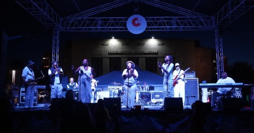 A live music performance on stage at night, featuring a group of singers and musicians in white outfits.