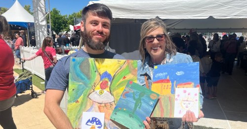A man and a woman smile while holding colorful paintings at an outdoor event with tents in the background.