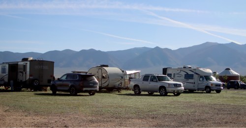 A row of RVs and trucks parked in a grassy area with mountains in the background under a clear blue sky.