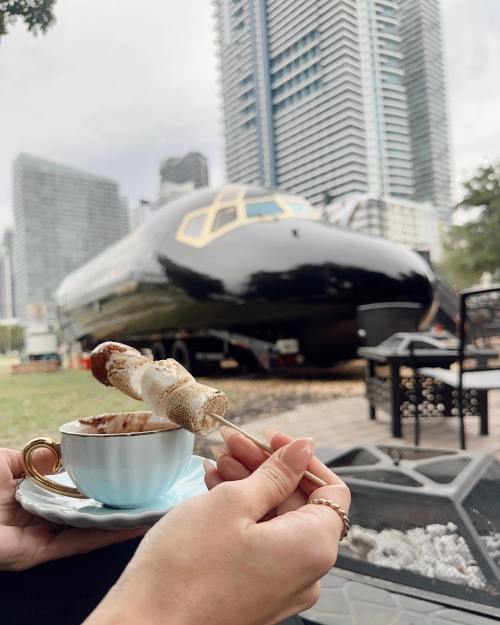 A person holds a cup of coffee and a skewer while sitting in front of a black airplane in an urban setting.
