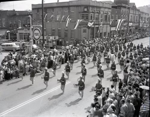 A black and white photo of a parade with marching bands and crowds lining the street, flags hanging above.
