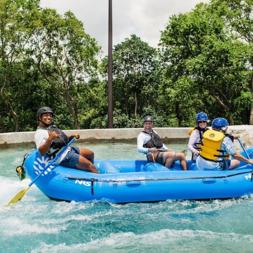 A group of four people in a blue raft paddling through a river, surrounded by lush greenery and a sunny sky.