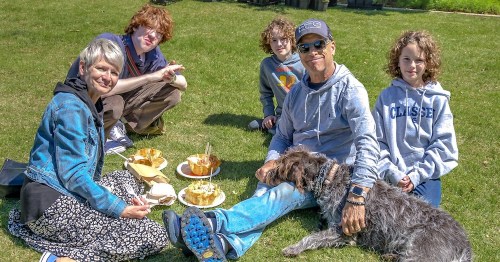 A group of five people and a dog enjoy a picnic on the grass, sharing food and smiling together.