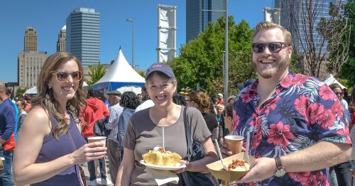 Three smiling people at an outdoor event, holding food and drinks, with a city skyline in the background.