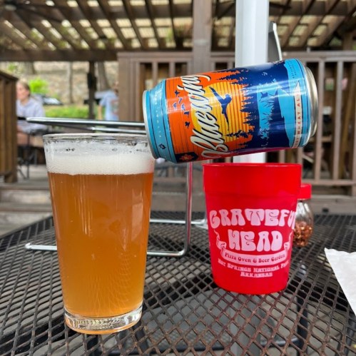 A glass of beer sits on a table next to a colorful beer can, with a red cup in the background at an outdoor setting.