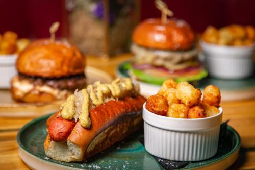 A hot dog topped with mustard, served with a side of crispy tater tots, with burgers in the background.