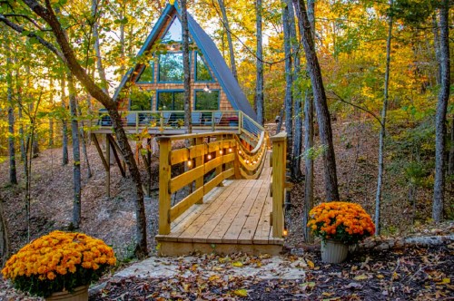 A modern A-frame cabin surrounded by autumn trees, with a wooden walkway and vibrant orange flowers in planters.