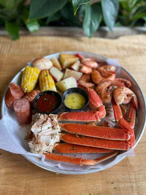 A seafood platter featuring crab legs, shrimp, corn, potatoes, sausage, and dipping sauces on a metal tray.