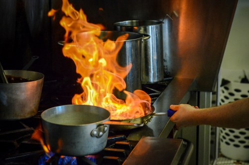 A chef's hand holds a pan over a stove, with flames erupting from the pan in a busy kitchen setting.