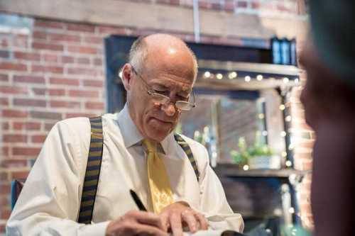 A man in a white shirt and yellow tie writes notes, wearing glasses and suspenders, in a cozy, brick-walled setting.