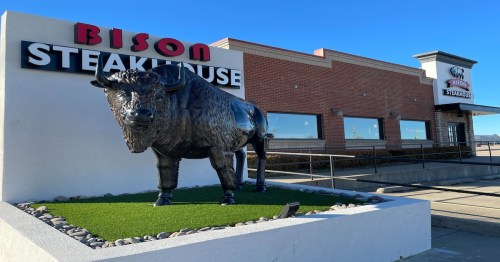 A large bison statue stands in front of the Bison Steakhouse, with a brick building and clear blue sky in the background.