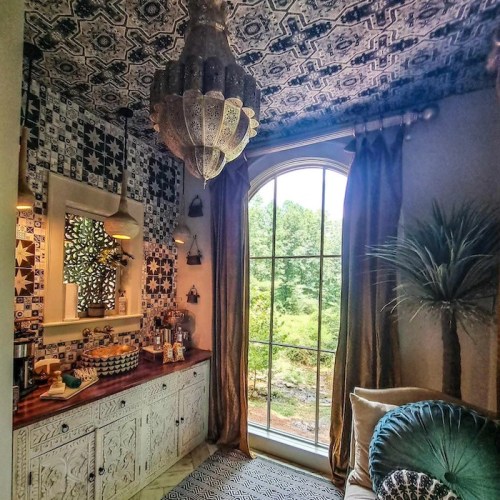 Cozy room with patterned ceiling, chandelier, decorative mirror, and a view of greenery through a large window.