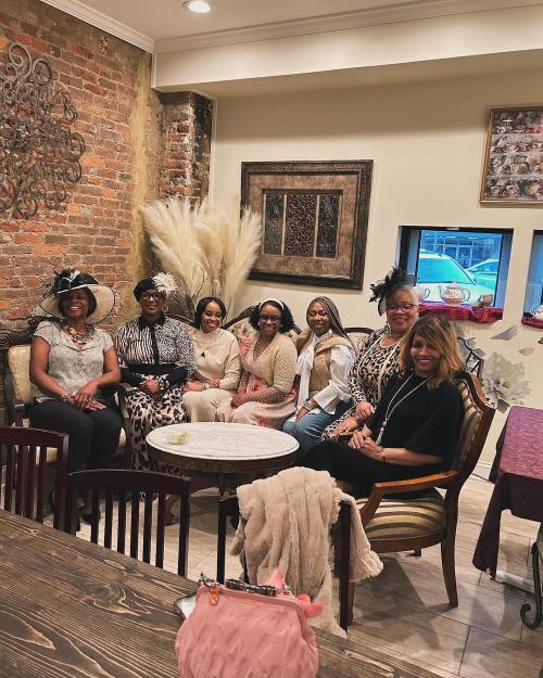 A group of seven women in stylish outfits sitting together in a cozy café with a brick wall backdrop.