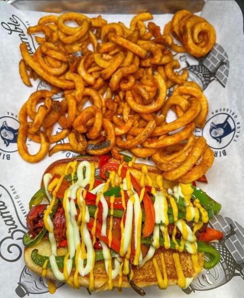 A loaded hot dog topped with grilled peppers and drizzled with mayonnaise, served with curly fries on the side.
