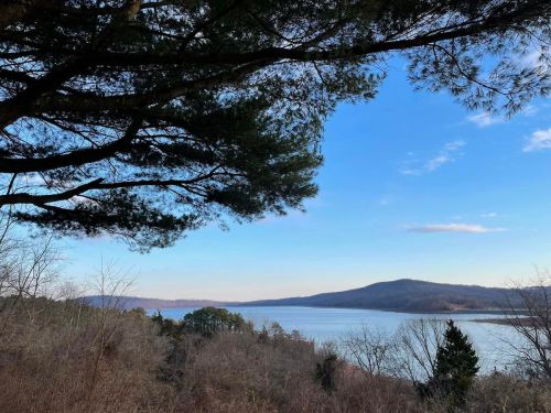 A serene view of a lake surrounded by trees and hills under a clear blue sky.